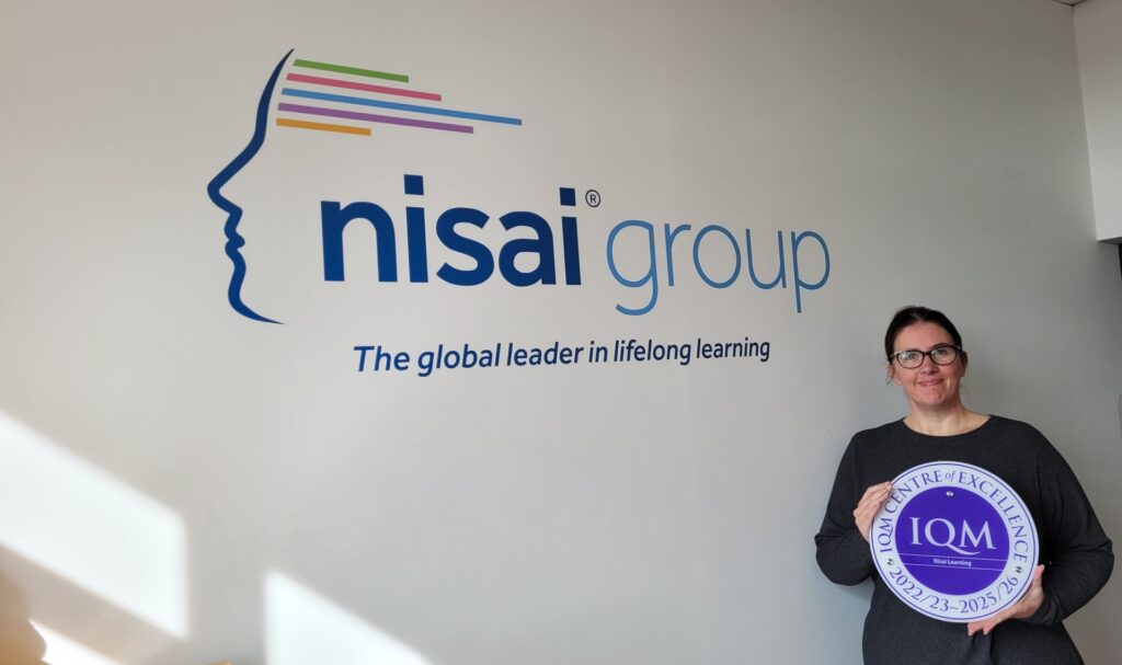 Ms. Victoria Reaney, Chief Operating Officer of Nisai, holding IQM “Centre of Excellence” Award while standing next to Nisai Group logo