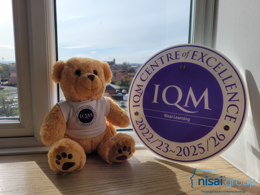 The image shows Dexter the Bear sitting on the windowpane with Nisai's IQM "Centre of Excellence" Award (2022/23 ~ 2025/26)